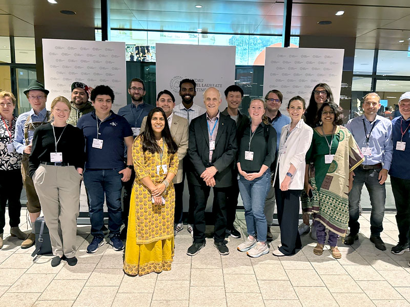 A diverse group of smiling people pose together in front of a board promoting the Lindau Foundation. 